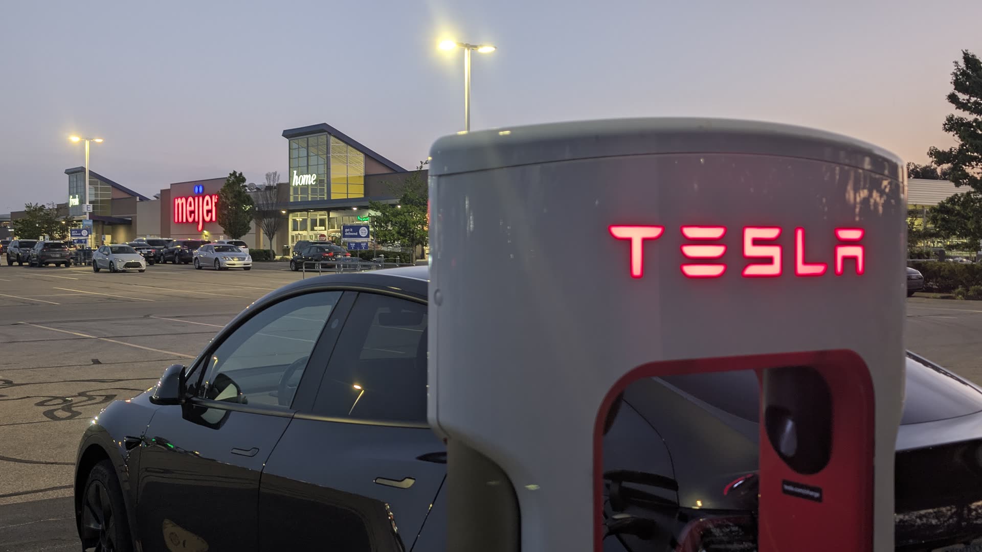 A Tesla Supercharger in a shopping-center parking lot at dusk, with a store and cars nearby