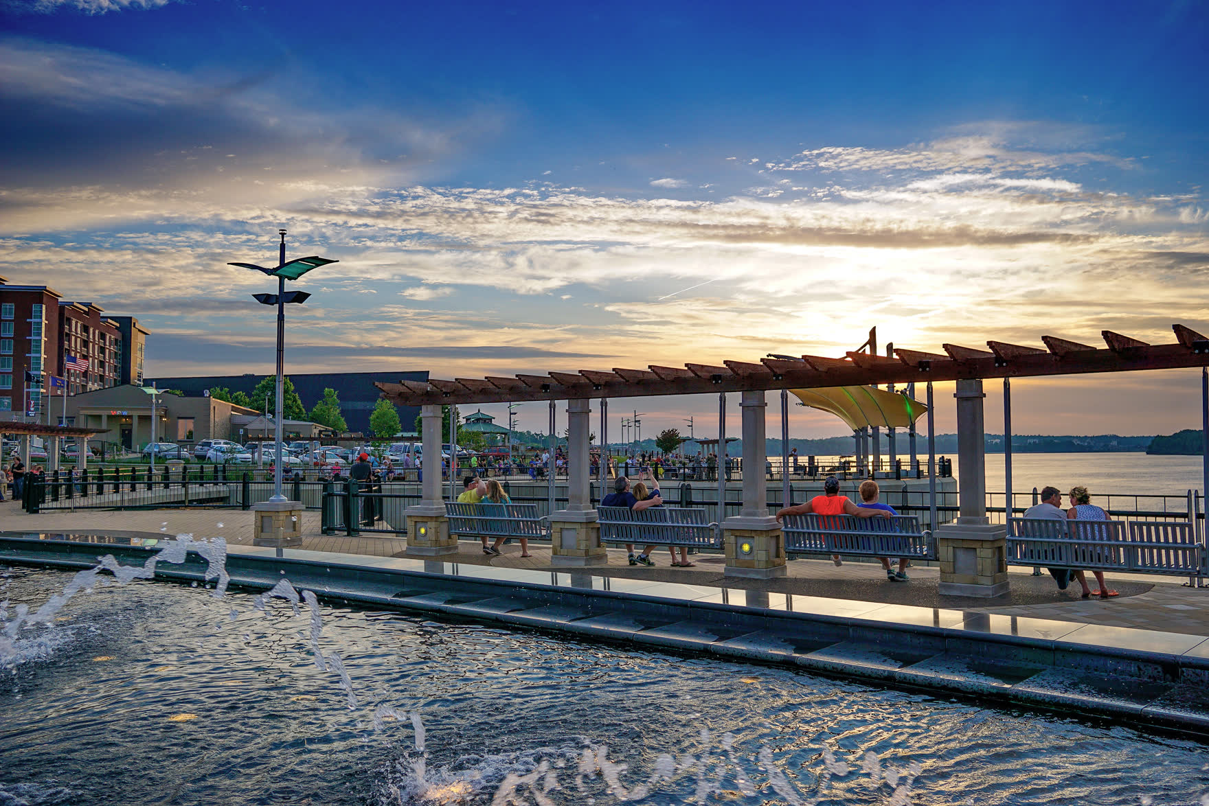 People relaxing along the Owensboro riverfront on the Ohio River at sunset, with fountains and a waterfront pavilion